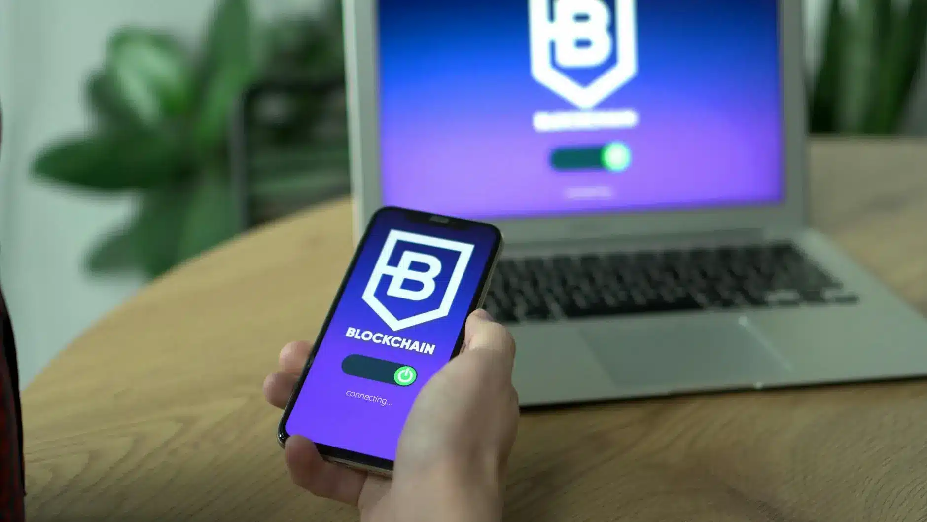 A user holding a smartphone displaying a blockchain app with a laptop in the background.