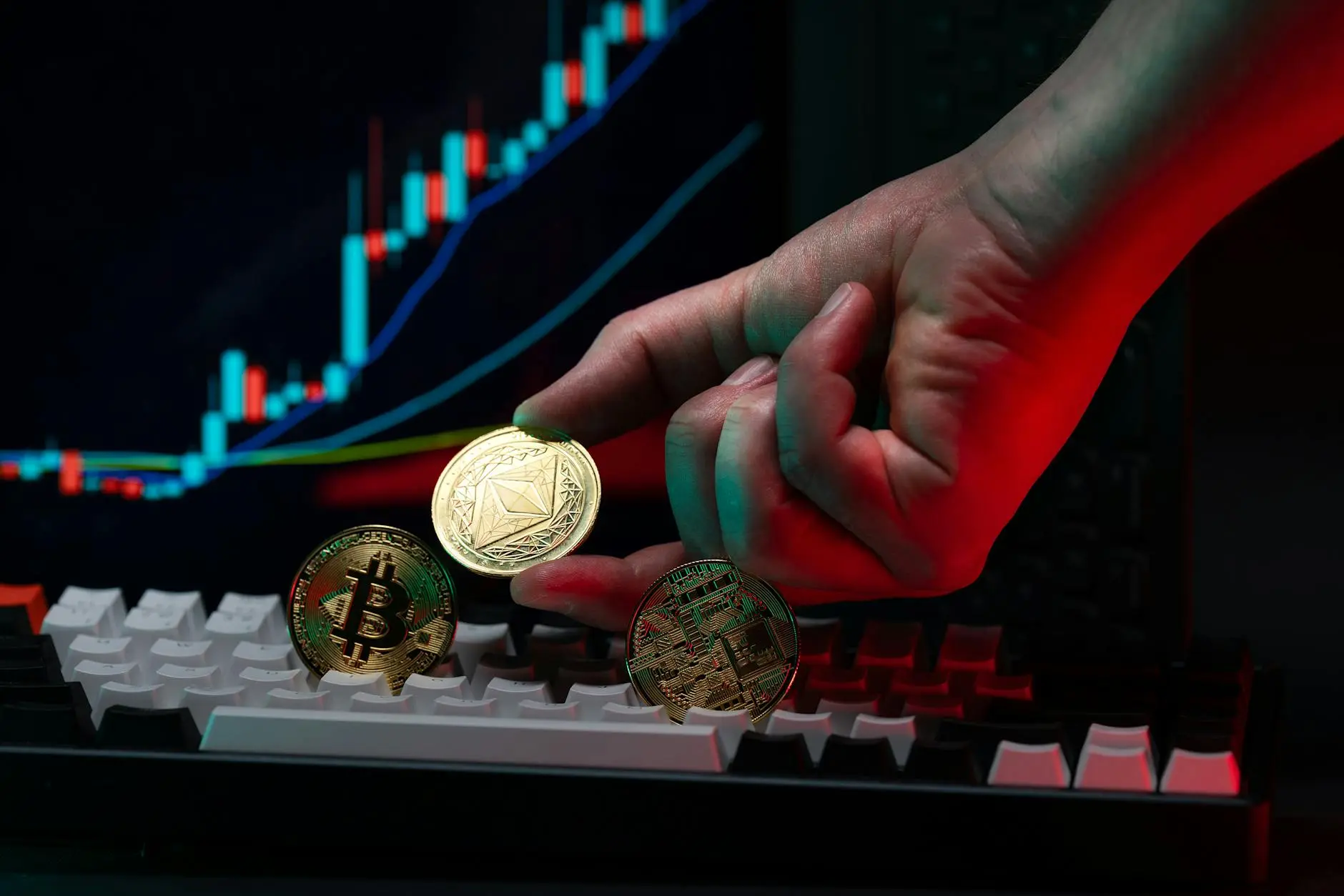 A hand holding a gold Ethereum coin above a Bitcoin coin on a keyboard with a trading graph background.