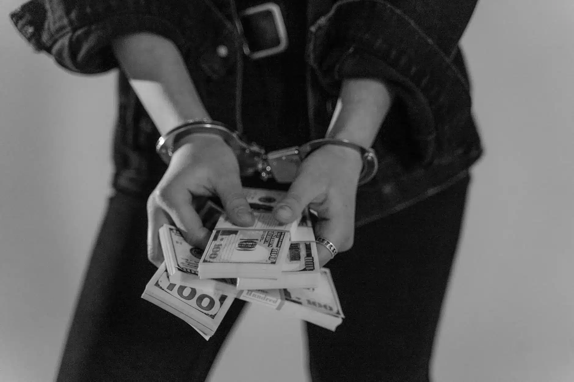 Hands in handcuffs holding stacks of hundred-dollar bills.