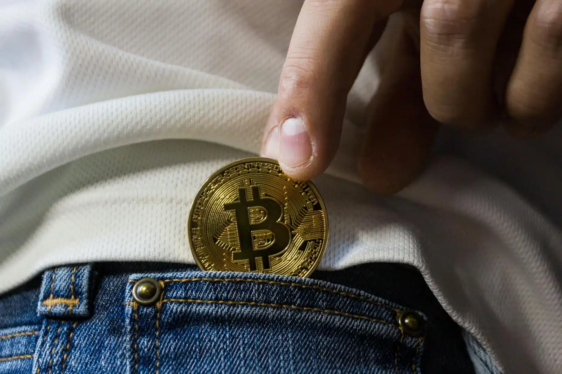 A hand holding a golden Bitcoin near the pocket of blue jeans.