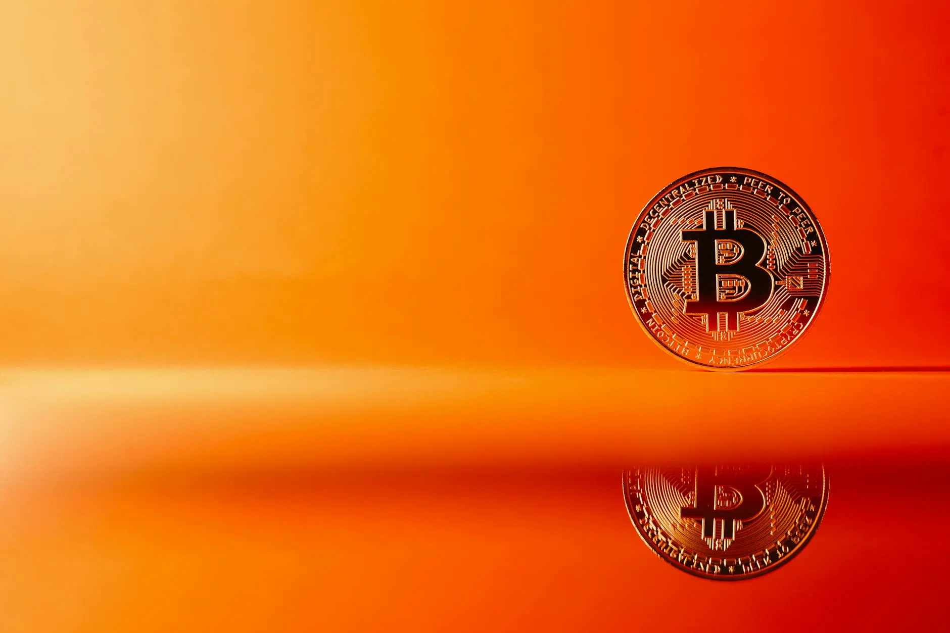 A Bitcoin coin on an orange background with its reflection visible.