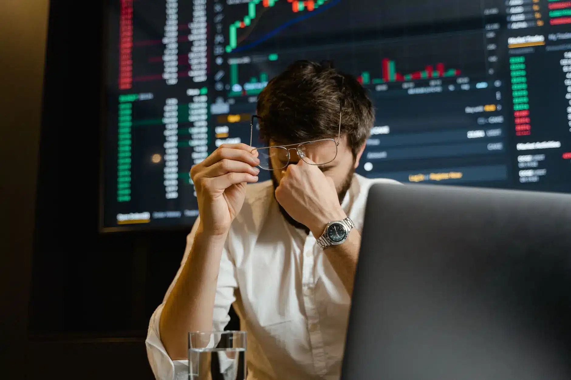 A stressed trader holding his glasses and resting his forehead on his hand in front of market charts.