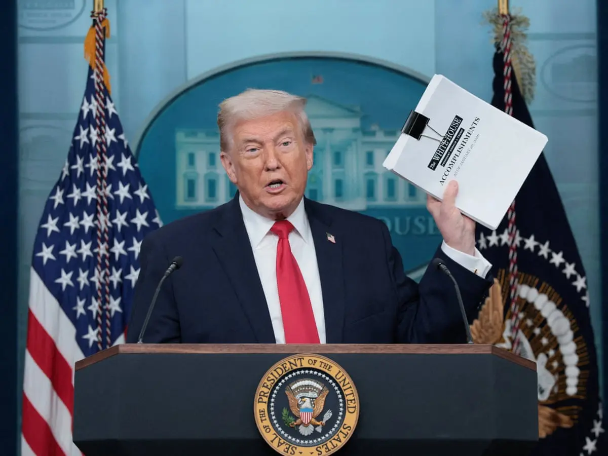 Donald Trump at a press conference holding a binder labeled 'Accomplishments'.