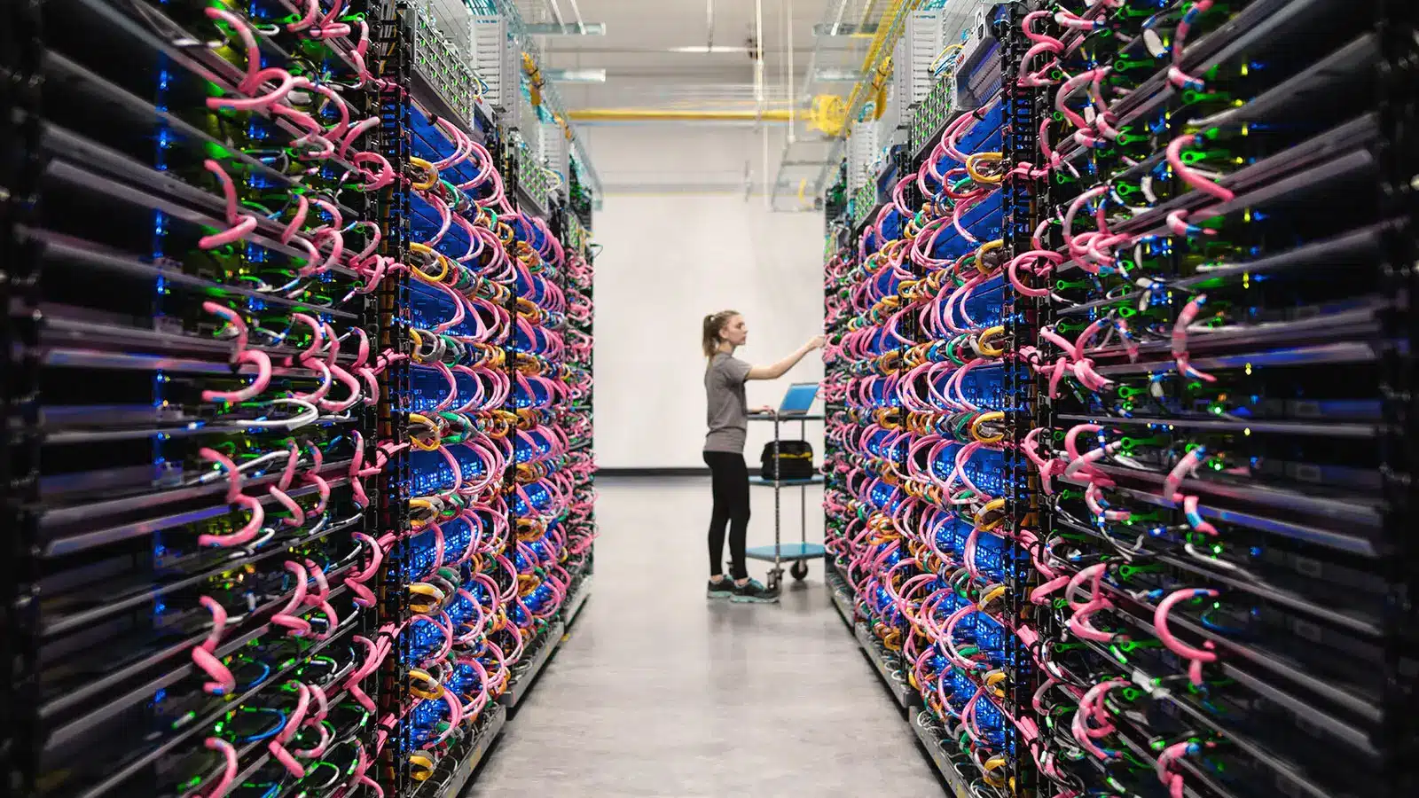 A technician working in a high-density datacenter aisle with server racks and colorful pink and green cabling.
