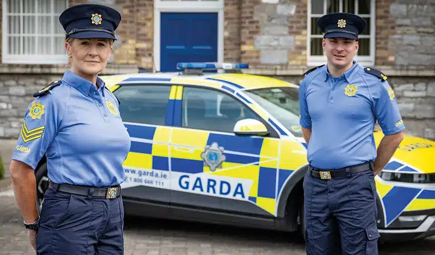 Two An Garda Síochána police officers in light blue shirts and dark caps standing in front of a marked police car