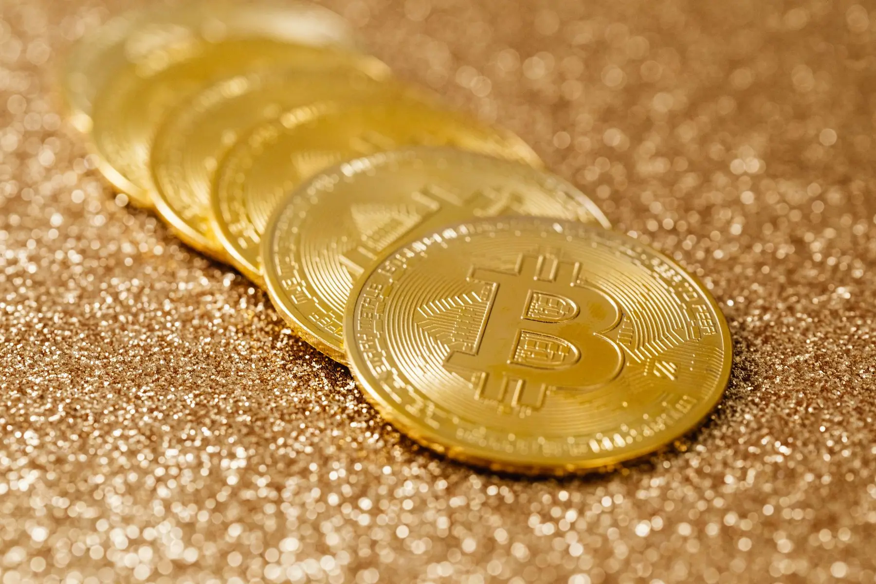 A row of gold-colored Bitcoin coins resting on a sparkling gold glitter background