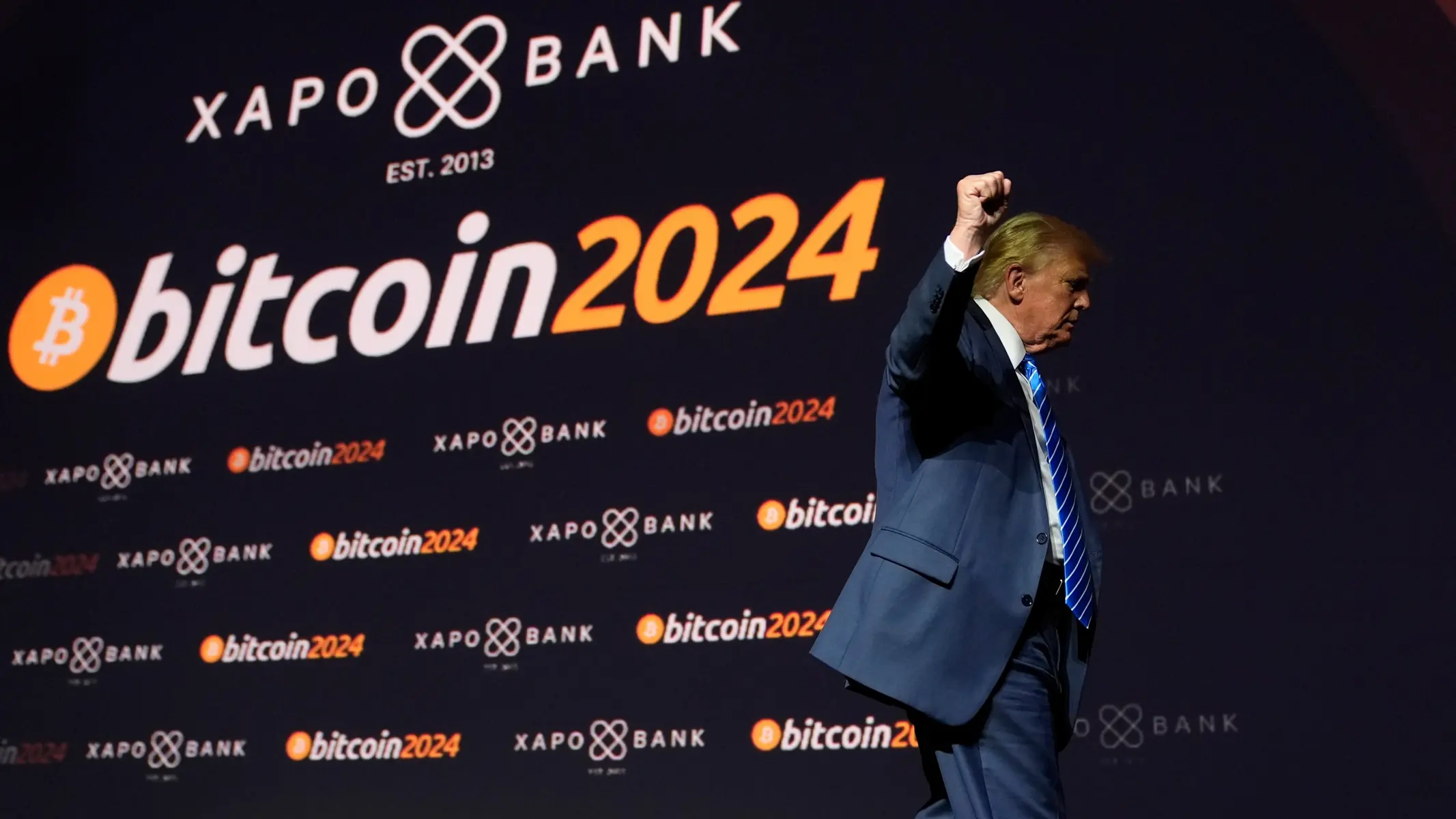 Donald Trump in a blue suit with a raised fist standing in front of a Bitcoin 2024 backdrop