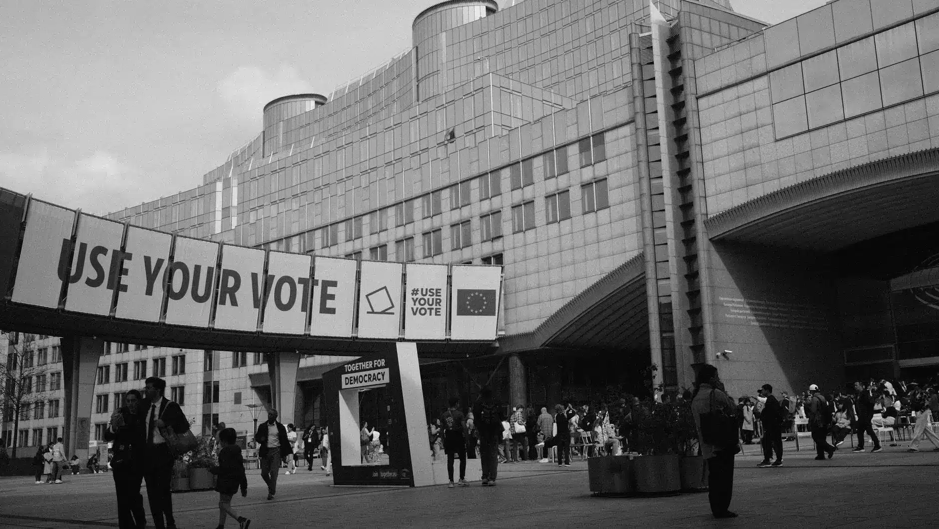 Black and white wide shot of the Berlaymont building in Brussels with a 'Use Your Vote' banner in the foreground