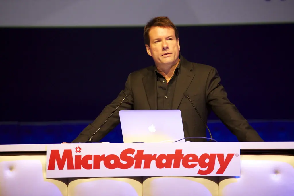 Michael Saylor speaking at a podium with a MicroStrategy logo and a laptop
