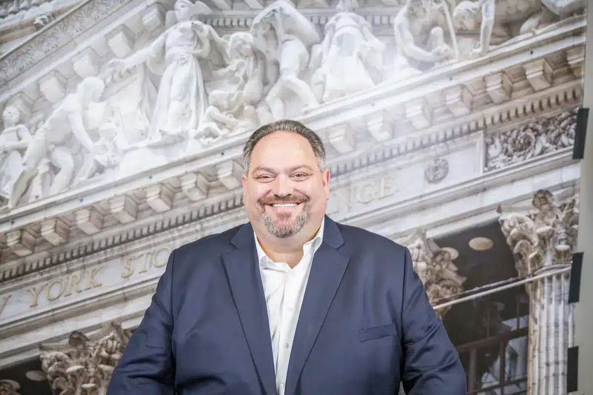 Portrait of Milton Todd Ault III smiling in a navy blue blazer and white shirt against a backdrop of the New York Stock Exchange.