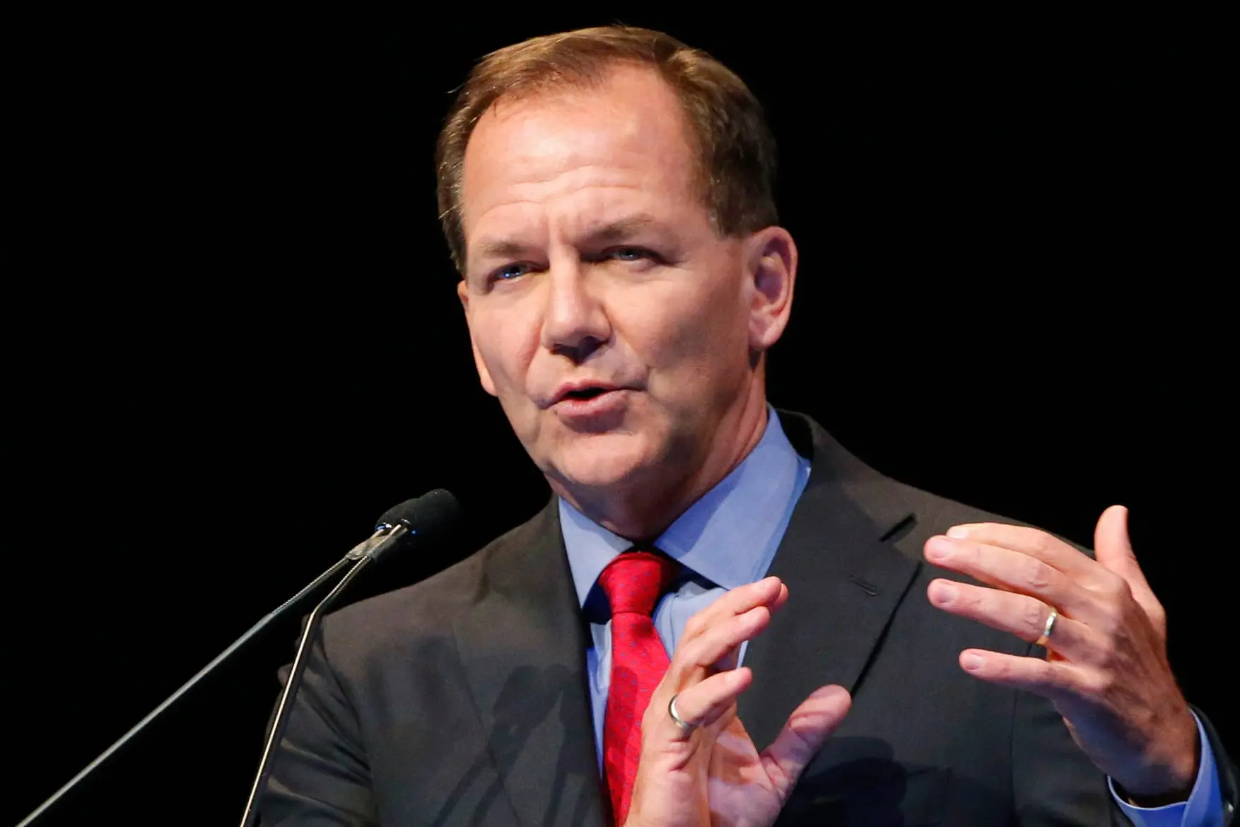 Paul Tudor Jones speaking at a podium with a microphone, wearing a dark suit and red tie