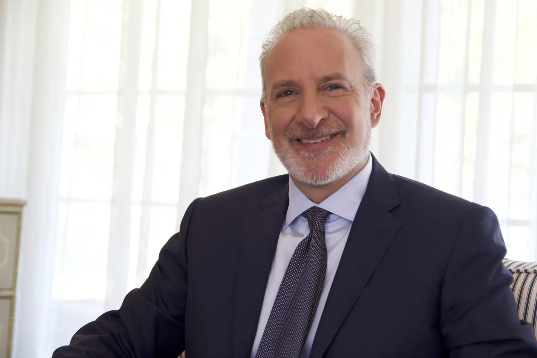 Portrait of Peter Schiff wearing a dark suit and patterned tie, smiling against a bright window background.
