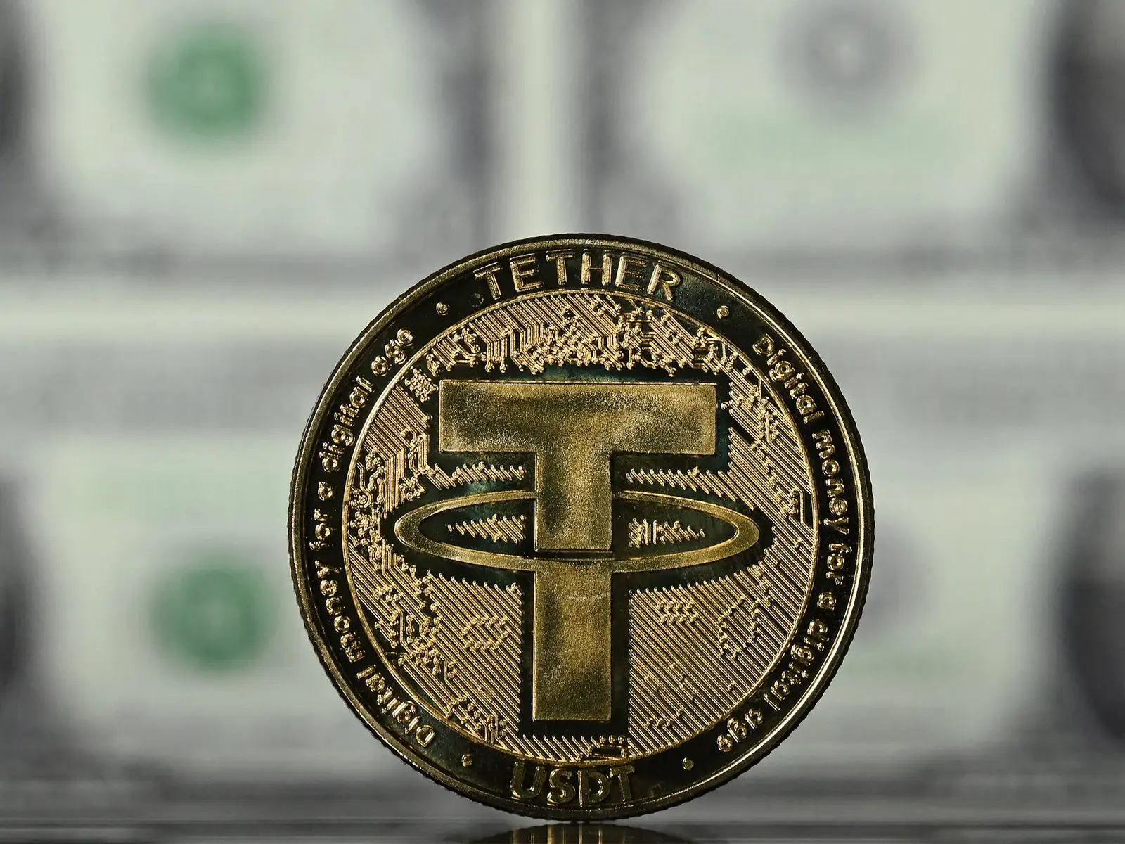 A gold-colored physical coin featuring the Tether USDT logo and text, set against a blurred background of US dollars.